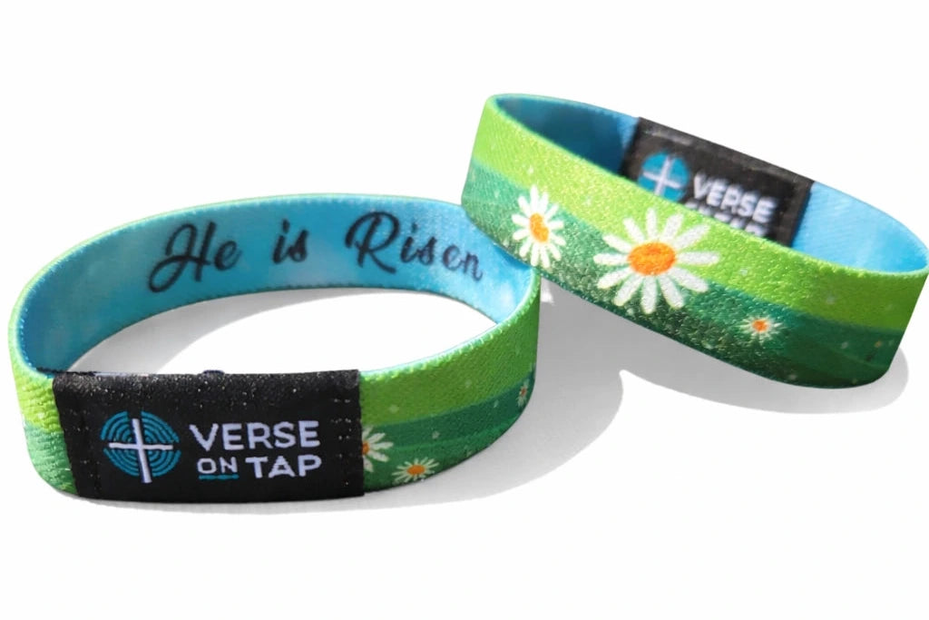 Elastic Christian wristband featuring green meadow with white daisies and blue interior band. “He is Risen” text with Verse on Tap logo tag visible.