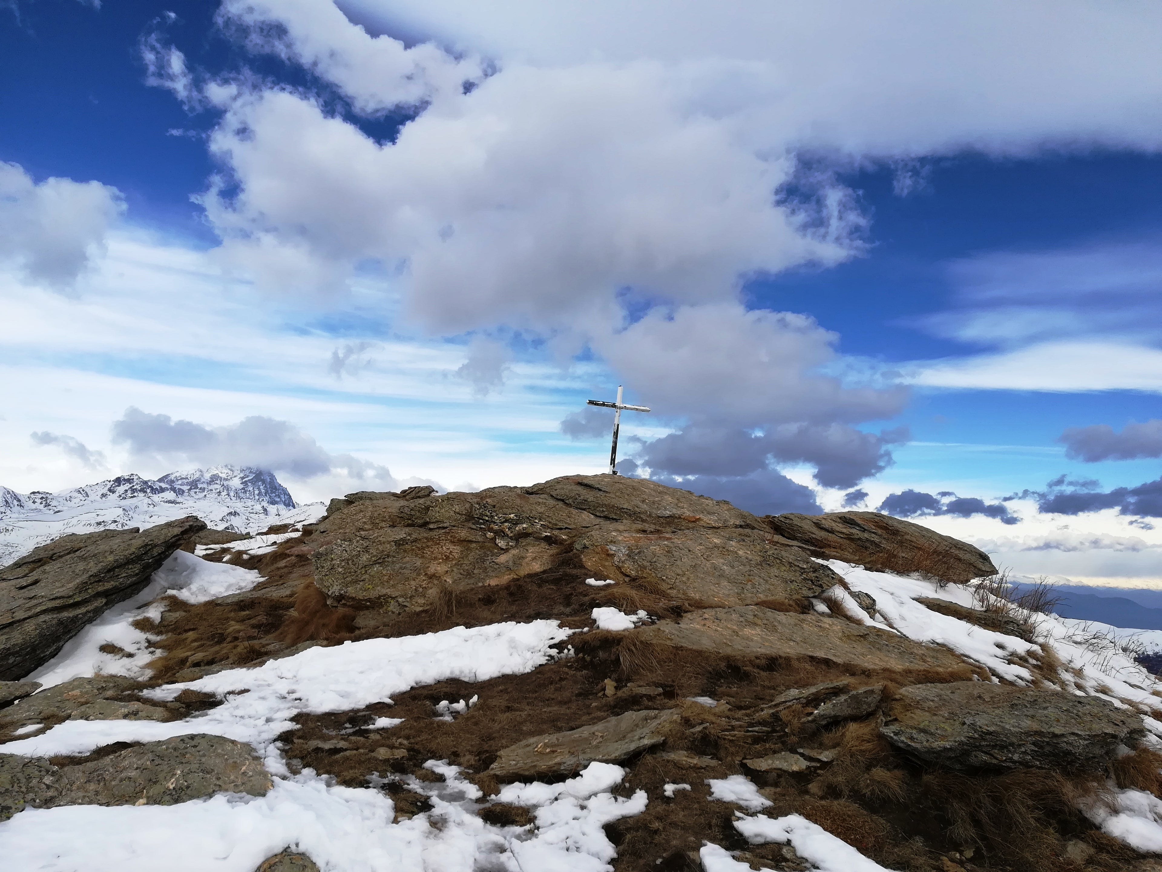 Wooden cross on a snowy mountain peak under a blue, cloud-filled sky