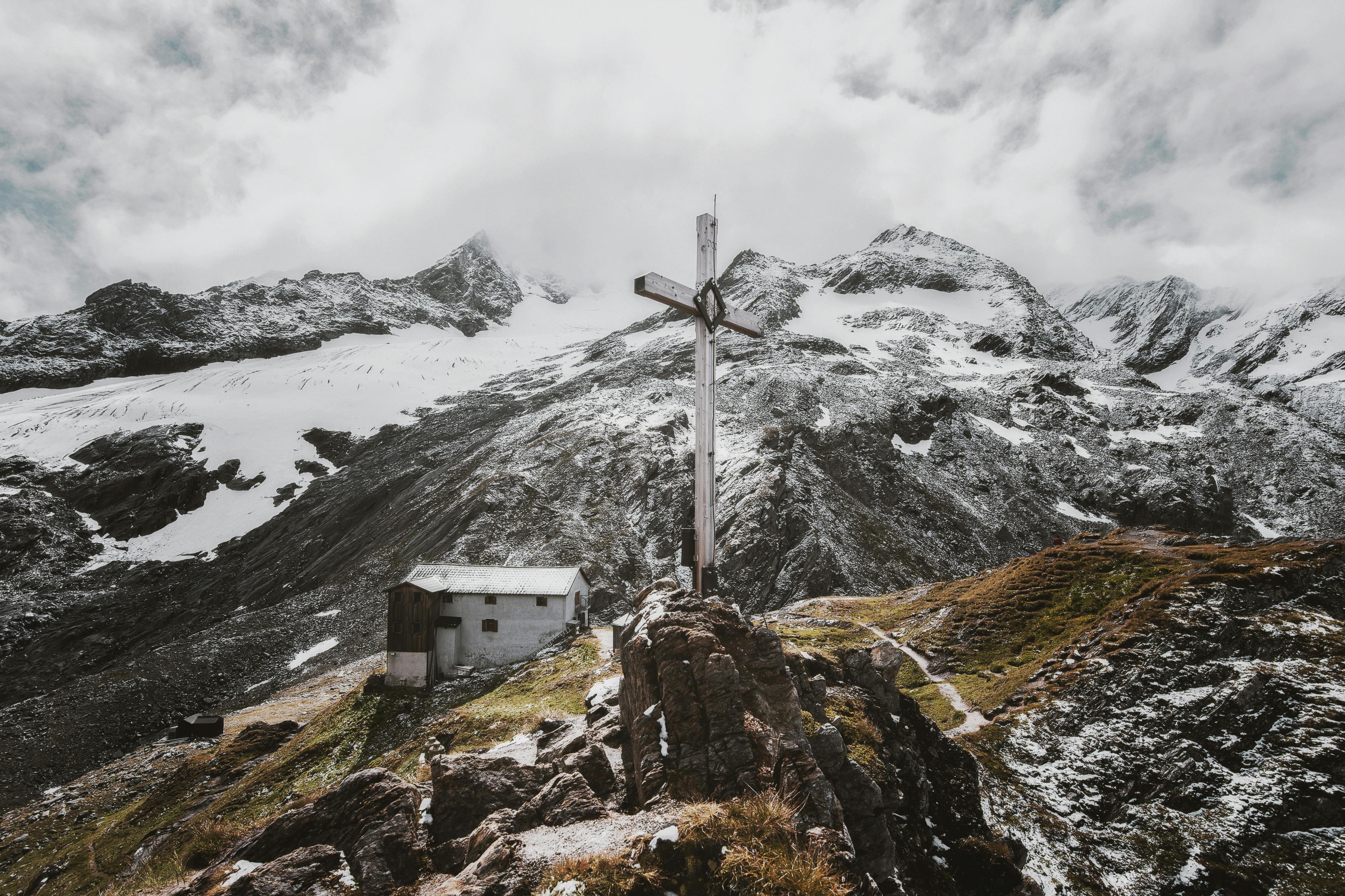 Wooden cross standing on a rocky mountain peak with snow-covered mountains and clouds in the background.